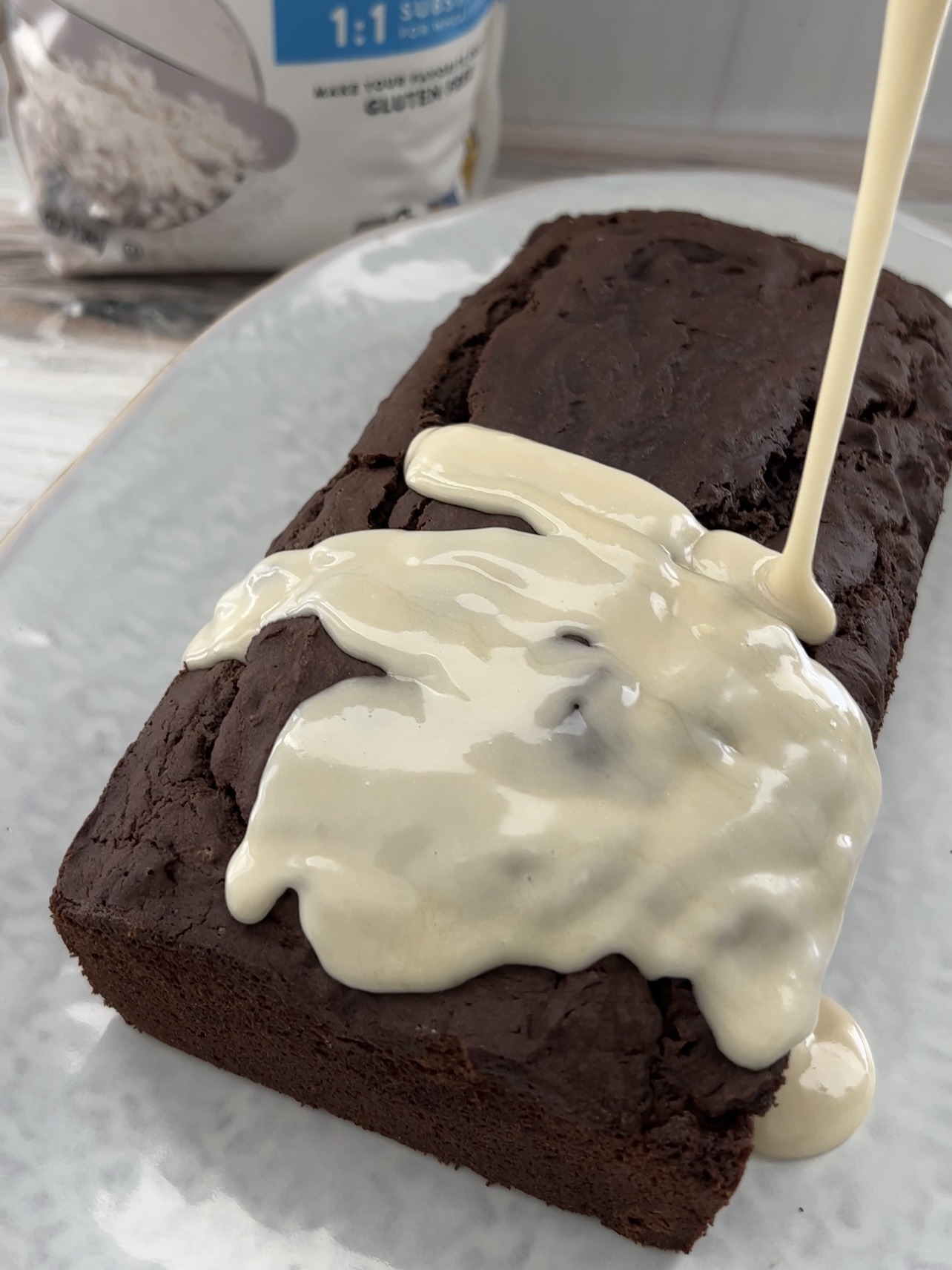 vegan chocolate cake on a white platter with white frosting being poured over top. 