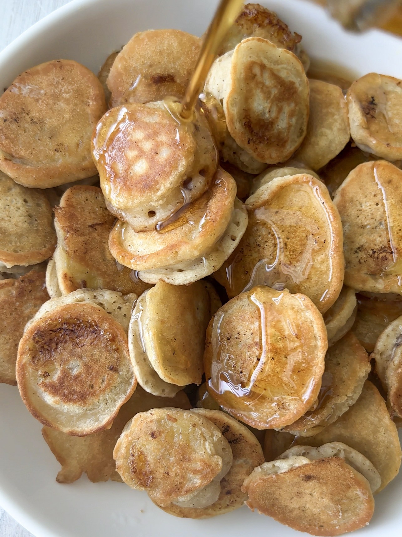 Mini pancakes on a white plate with maple syrup being poured on top. 