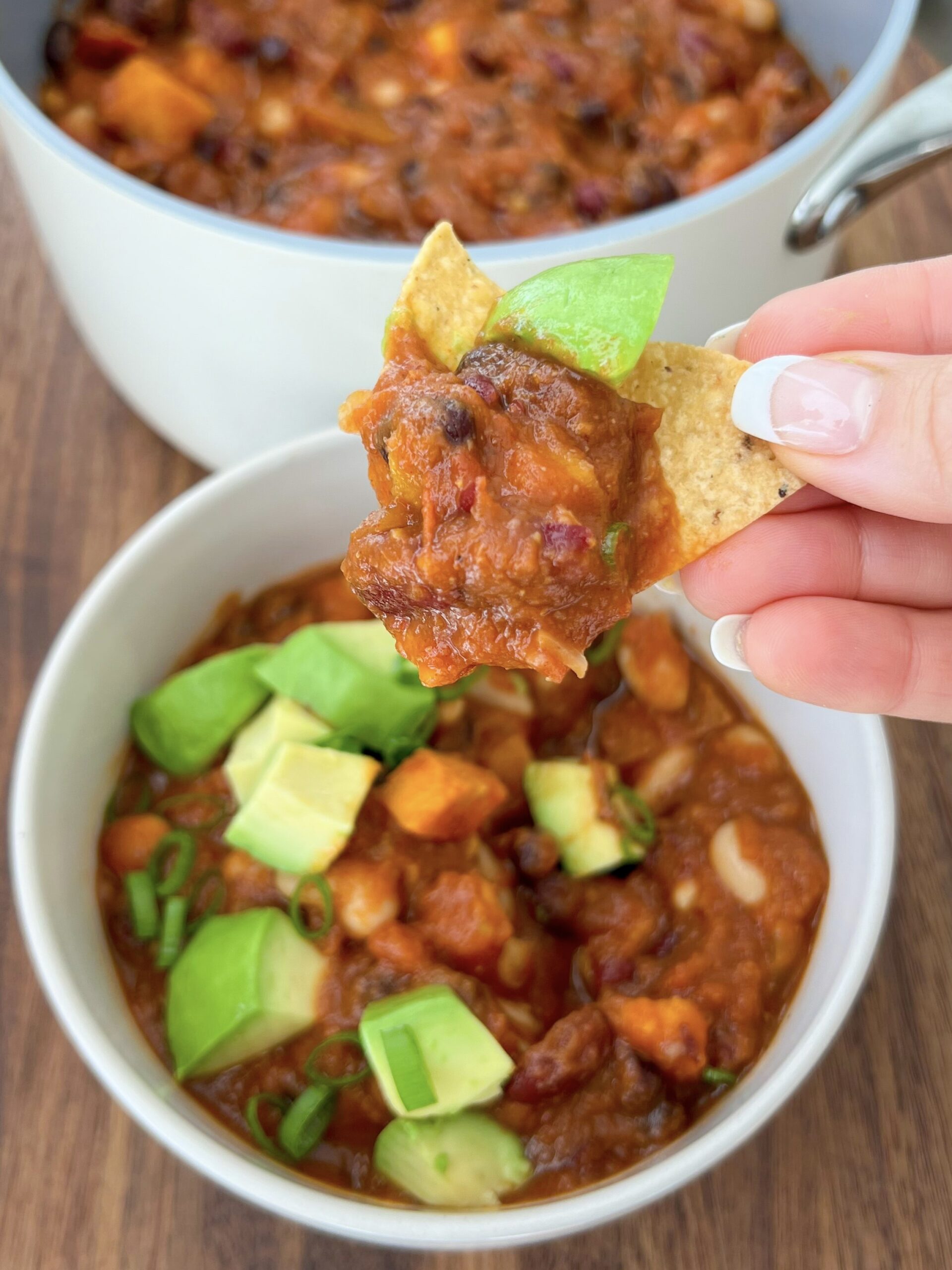 Hand with chip dipped in chili and bowl of chili in the background. 