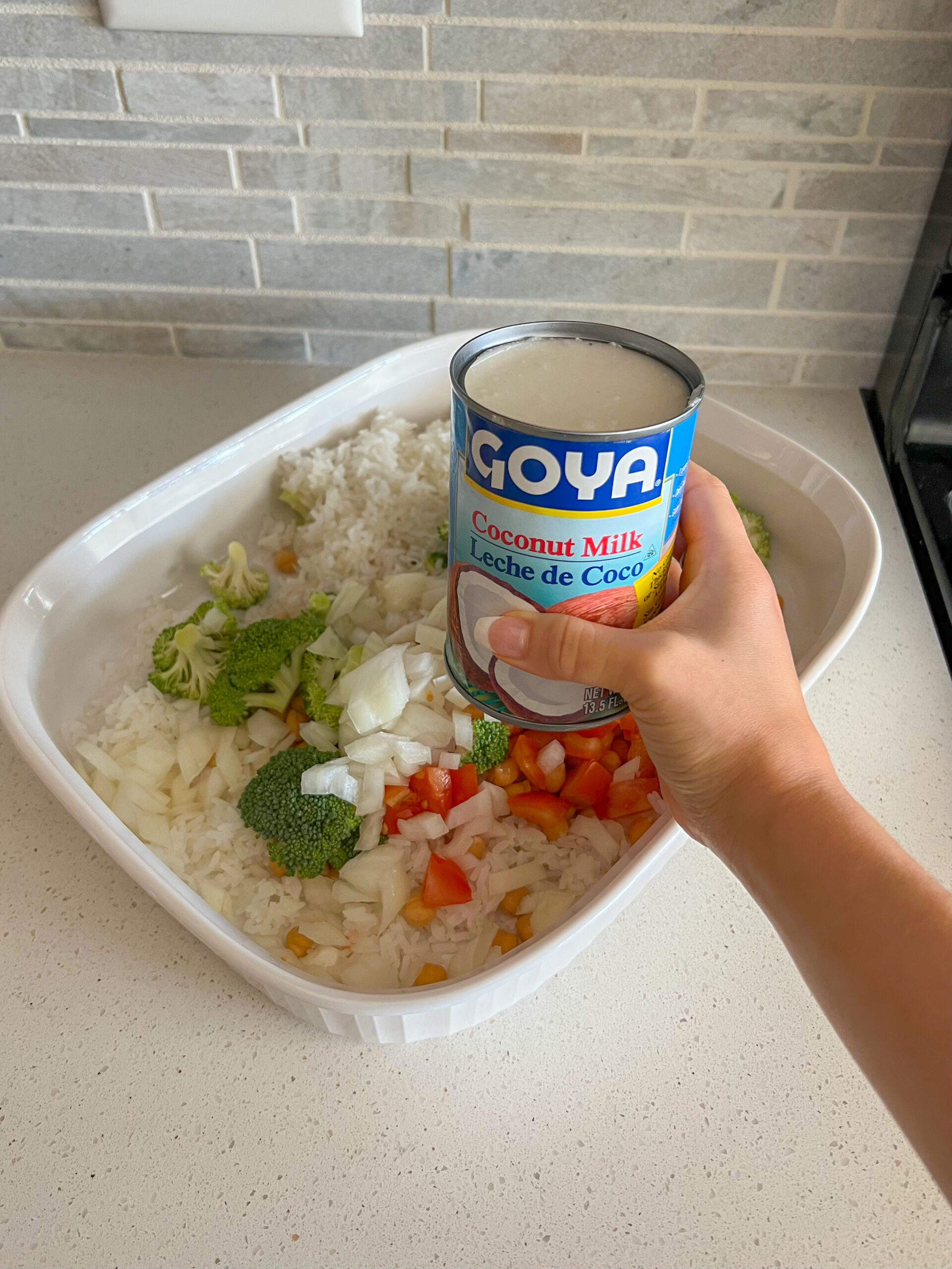 Rice and vegetables in a casserole dish with a hand holding a can of coconut milk above it. 