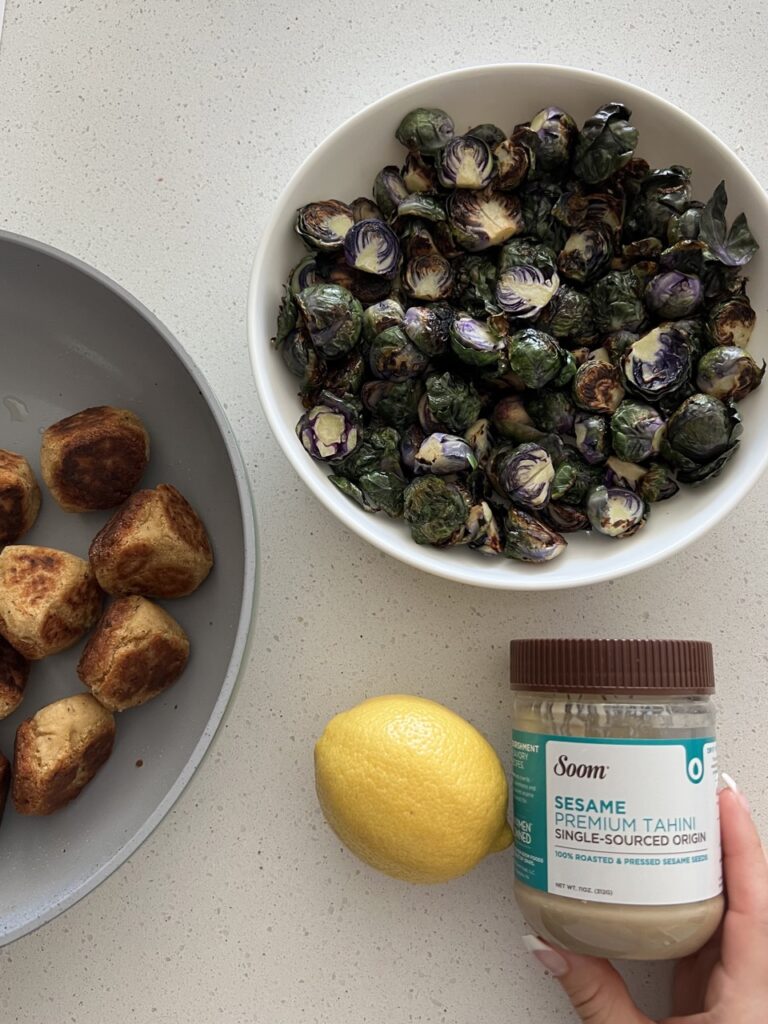 Two bowls with meatballs in one bowl and Brussels sprouts in the other along with a lemon and tahini. 