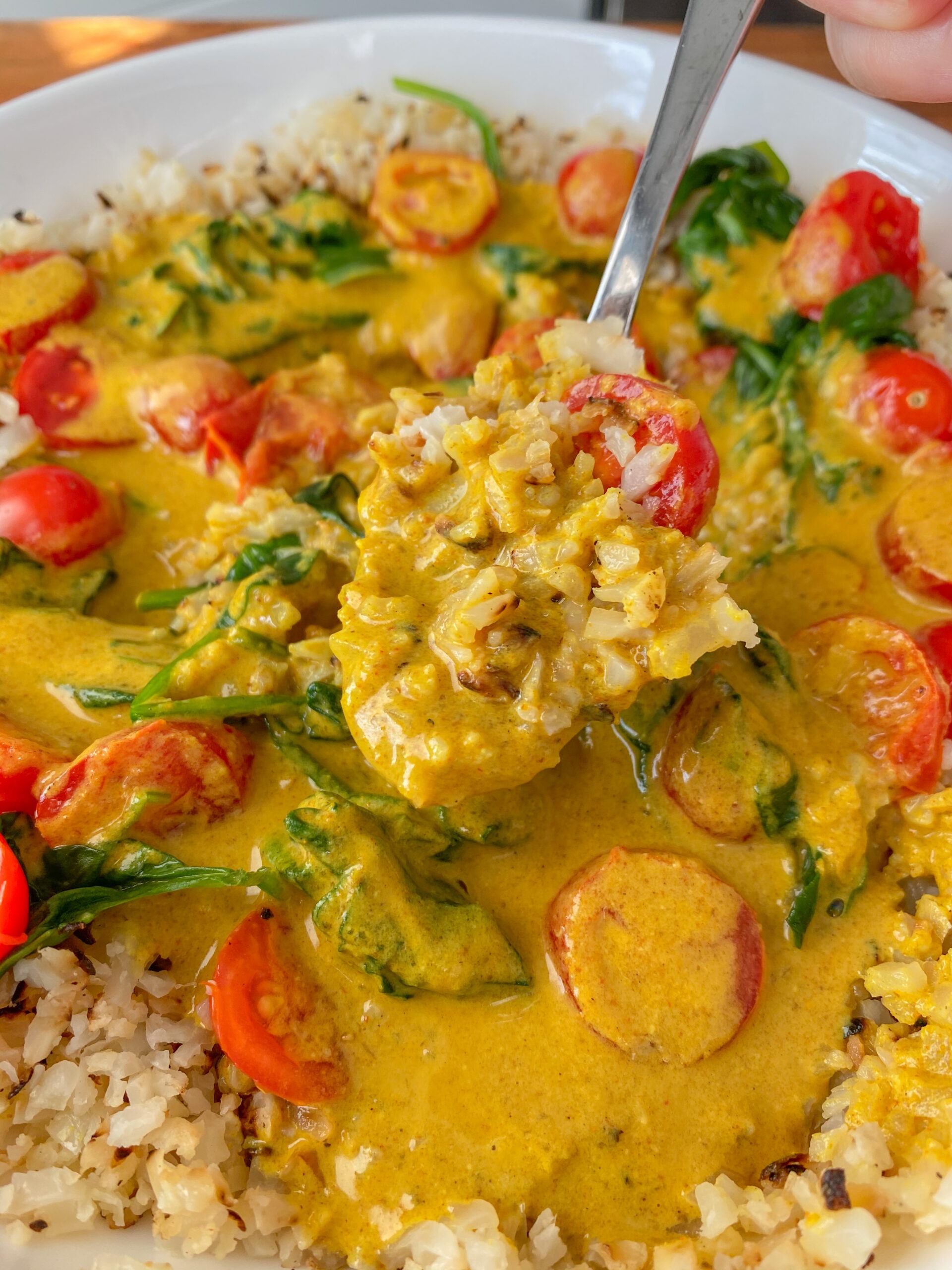 Bowl of coconut curry with tomatoes and spinach with a hand holding a spoonful of it. 