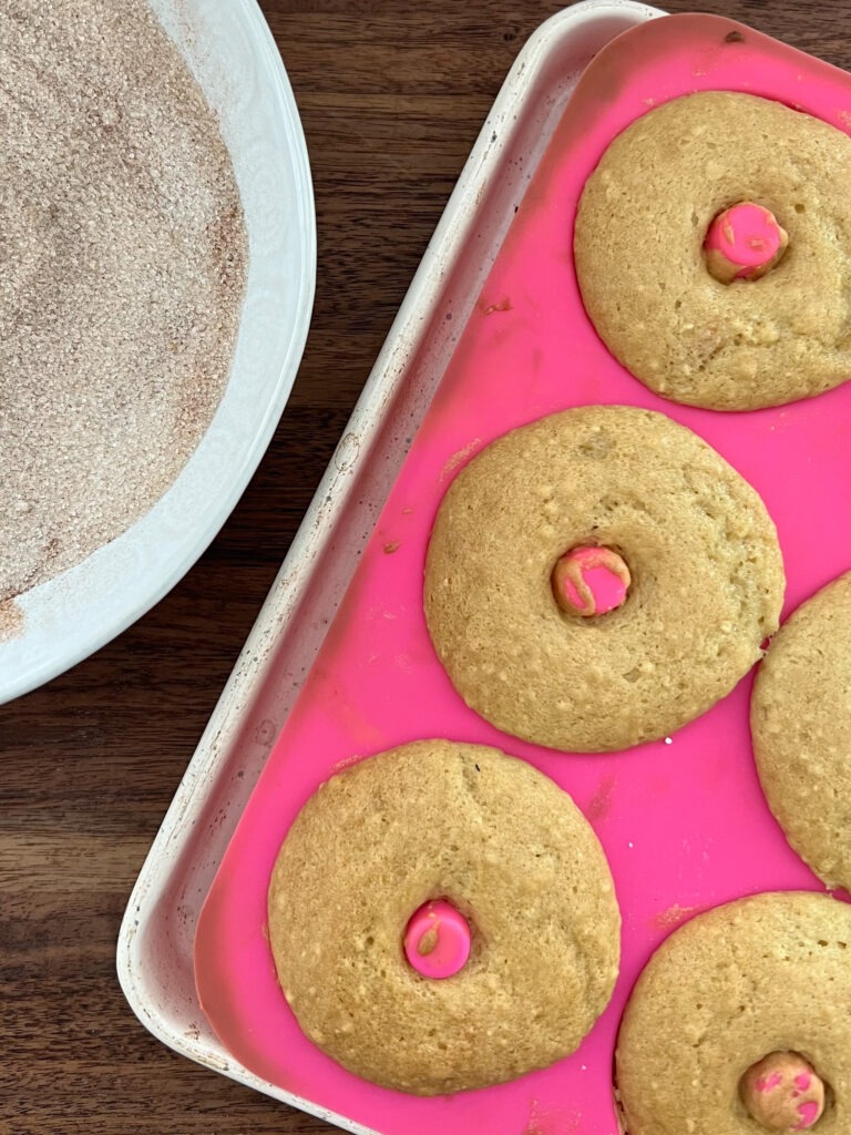 Donuts baked and in donut mold next to a bowl of cinnamon and sugar. 