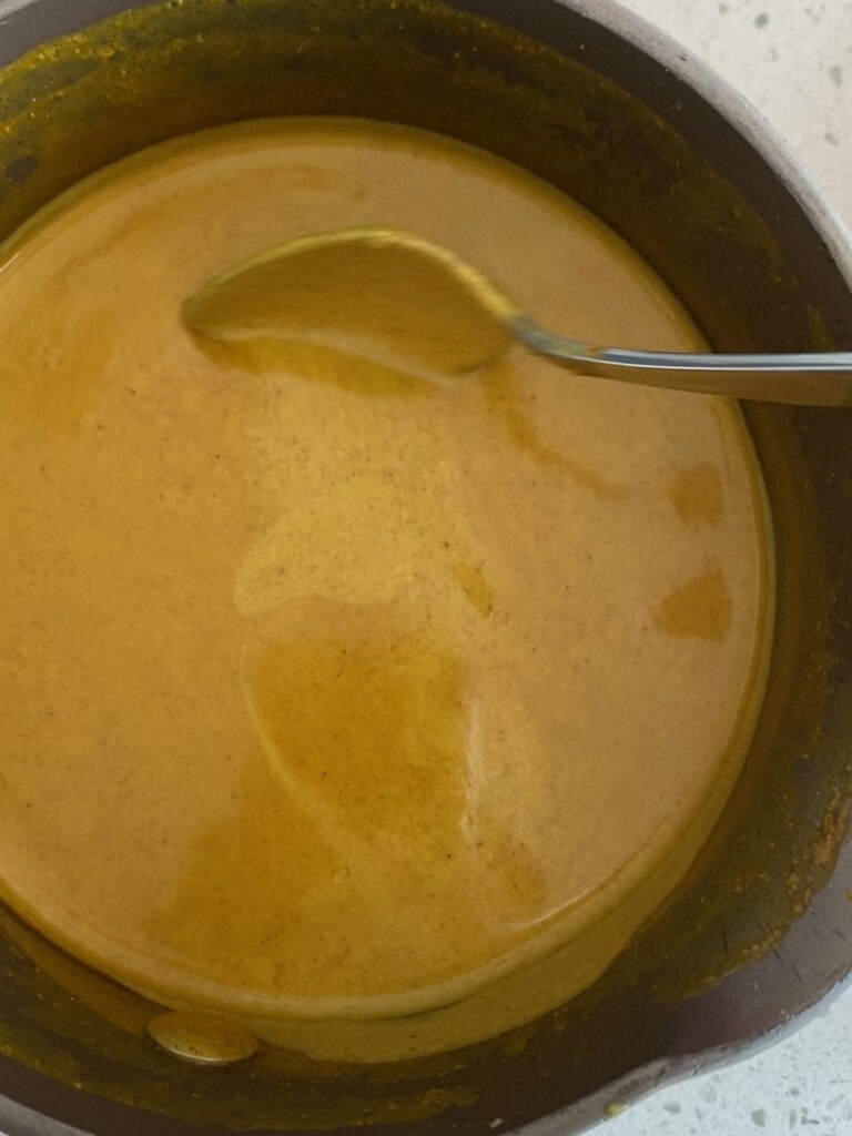 Coconut curry sauce in a saucepan, being stirred with a metal spoon. 